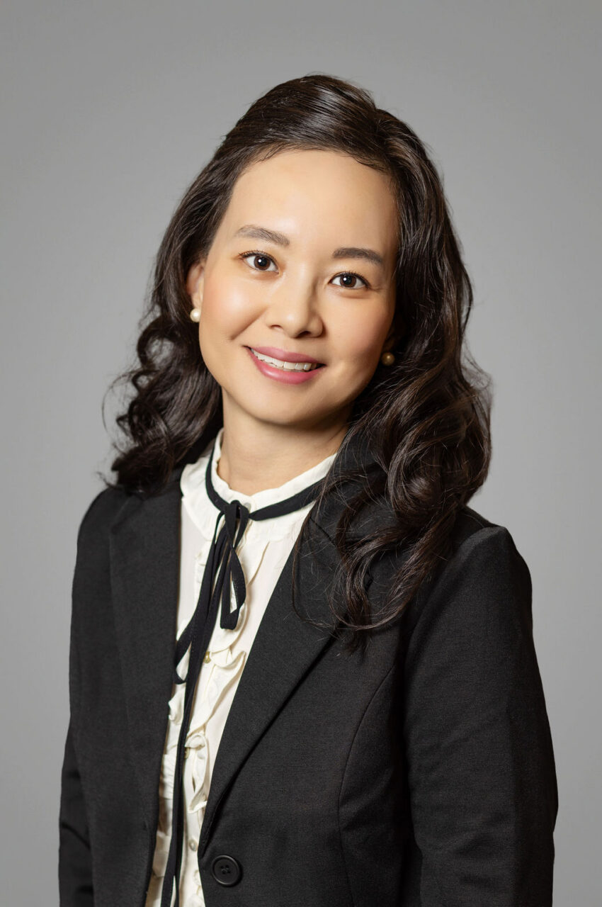 A professional headshot of an East Asian woman with long, dark wavy hair, wearing a black blazer over a patterned white blouse featuring a black tie neck accent, posed against a neutral gray background.